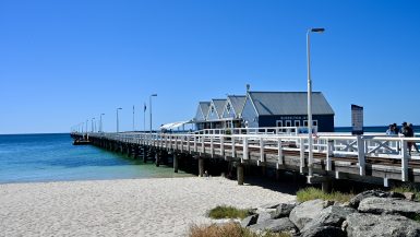 View across golden sand to a white wooden pier running from the right of the photo across to the horizon on the left. A row of wooden huts sits on the pier. Blue sky above. Turquoise sea visible to the left
