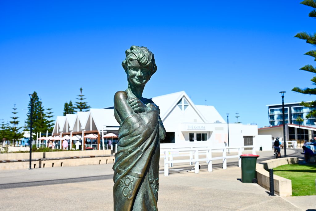 Brass statue of a girl holding towel around her with white building and blue sky behind her