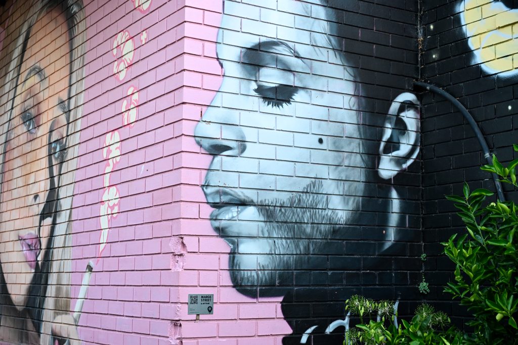 Corner of a brick building painted pink. To the right is a painting of the musician Prince. To the left is a painting of a lady with blond hair