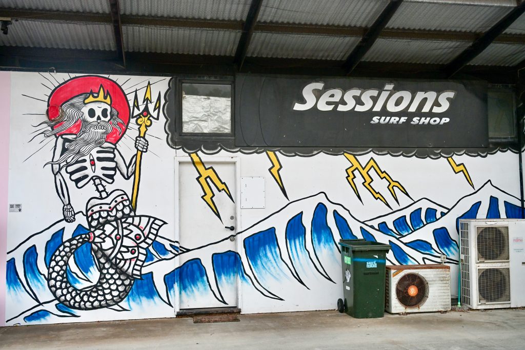 White wall mural with a painted blue wave at the bottom and skeleton mermaid figure to the left. Top right says Sessions in white on black