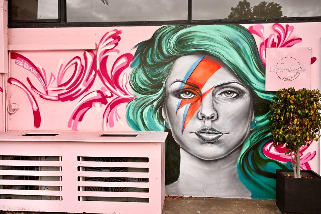 Pink mural on a wall. Features a lady to the right hand side with green hair and a David Bowie lightening bolt across her face