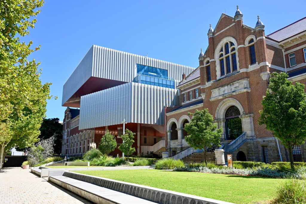 Looking at a modern building with 2 white block roofs sitting at juxtaposition to the left, An old red brick building to the right. Grass in front