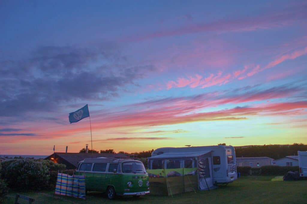Technicolour sunset sky with purples, pinks, oranges and golds over a campsite scene with green VW Camper can and white motorhome. Blue flag flying.