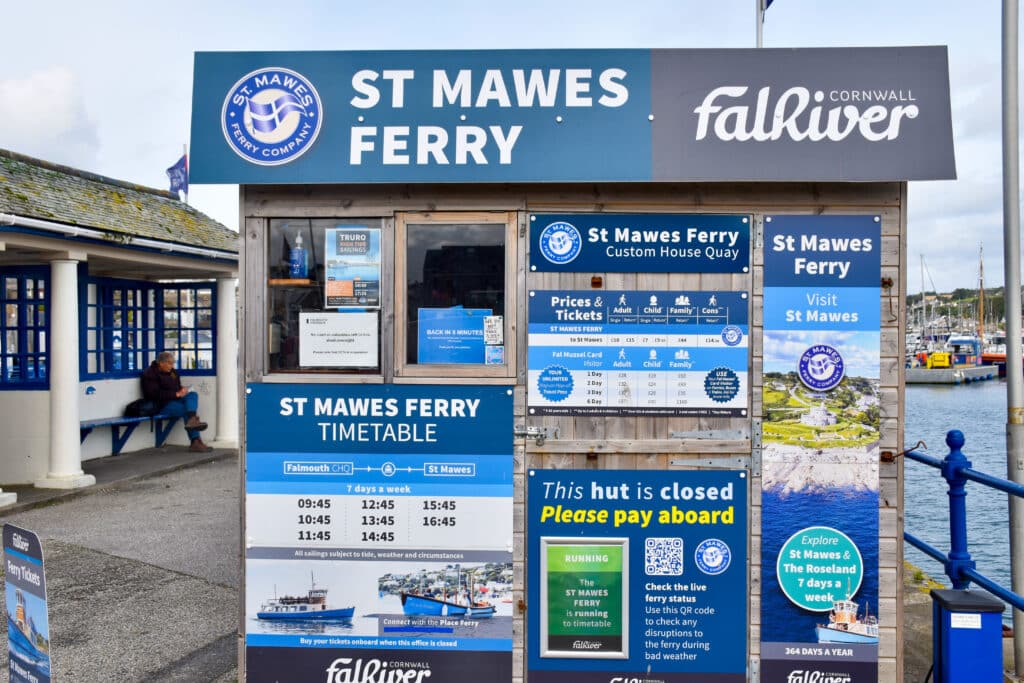 A hut fills the frame and has blue and white signs all over it advertising the St Mawes Ferry. It advertises the timetable, prices and tickets.