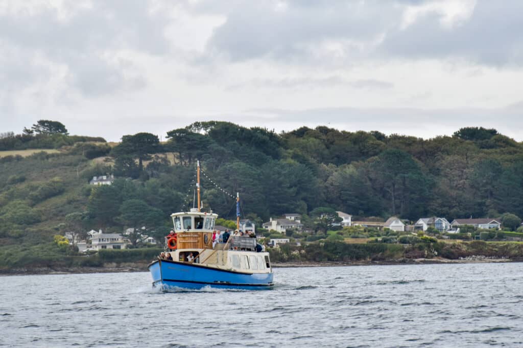 A blue hulled wooden ferry chugs towards the camera on a body of grey river. Grassy hillside with trees behind. Some houses lined up by the water. Grey sky above.