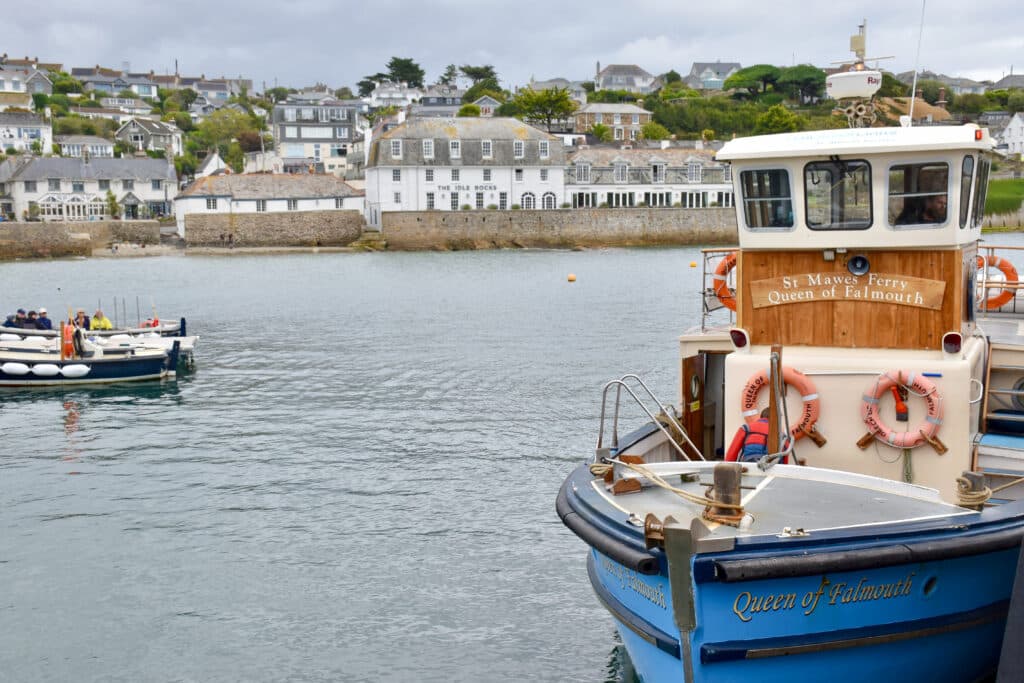 A wooden ferry boat with blue hull sits to the right hand side of the frame. Grey water of the harbour to the left. Rows of white cottages in the distance. the ferry says 'St Mawes Ferry Queen of Falmouth' on a wooden sign.