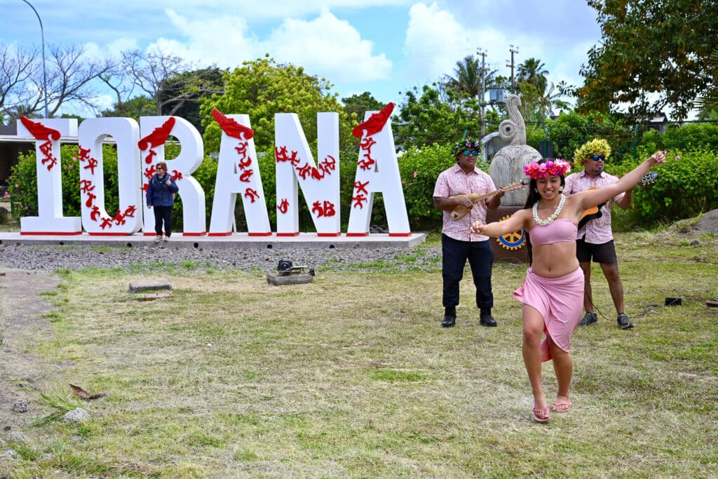 Looking across a stretch of pale green grass to large white letters spelling IORANA. To the right are two musicians with leaf wreaths on head and ukeleles and a dancer in pink outfit with pink flower garland