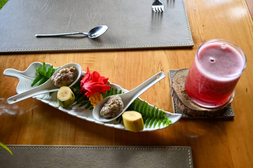 Looking down on a wooden table top with two place settings for dinner. In the middle is a white dish shaped like a palm leave with slices of banana on them, two spoons with scoops of ceviche and a tropical flower. A glass to the right has a red fruit juice in it