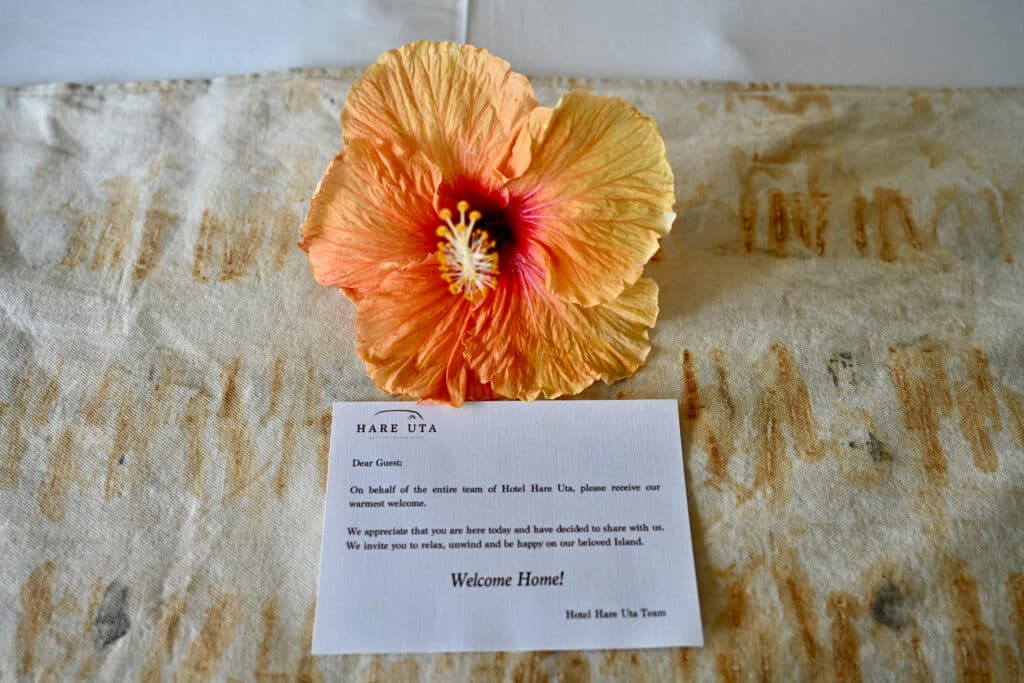Looking down on a white card with a welcome message from Hotel Hare Uta. Topped with an orange tropical flower