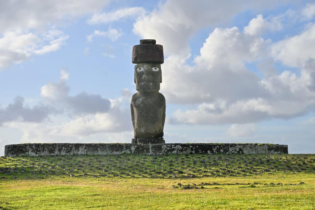 Grassy field in front. Wide stone platform above. Single carved stone statue stands in the middle of the platform facing the camera. It has white eyes and a red hat. Cloudy, pale blue sky behind.