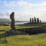 View across grass to a single stone human like statue to the left hand side. In the distance to the right is a platform with the remains of 5 stone figures on it. Coastline behind on the horizon. Pale blue sky above