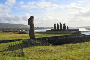 View across grass to a single stone human like statue to the left hand side. In the distance to the right is a platform with the remains of 5 stone figures on it. Coastline behind on the horizon. Pale blue sky above