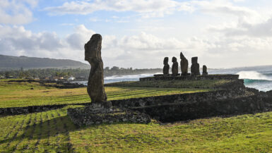 View across grass to a single stone human like statue to the left hand side. In the distance to the right is a platform with the remains of 5 stone figures on it. Coastline behind on the horizon. Pale blue sky above