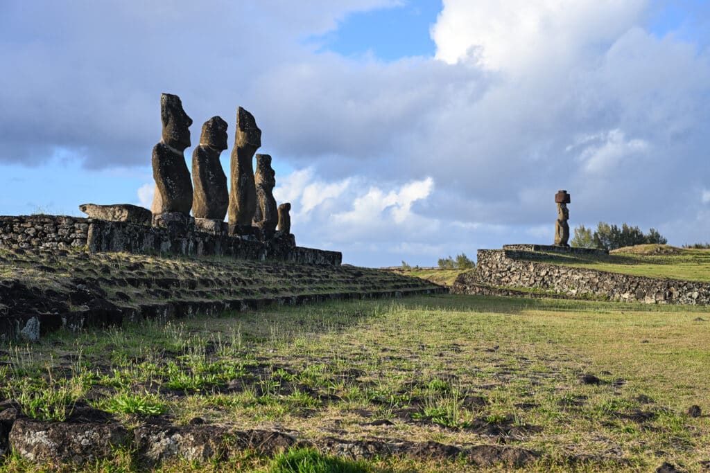 Platform of 5 stone statues to the left and a single statue to the right. All on stone platforms. Grassy fields below. Cloudy blue sky above.