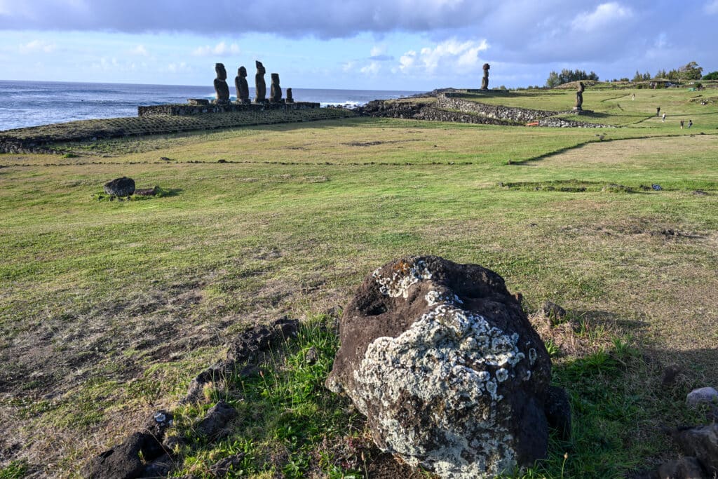 A weathered carved stone head lies in the bottom right of the frame, its chin covered in moss. Grass field reaches to the ocean at right hand side. Blue sky with bank of clouds above. A stone platform sits on edge of the grass supporting 5 stone figures. Two other stone figures are visible - one to the right on the coast with a hat on, and one lightly in land from that.