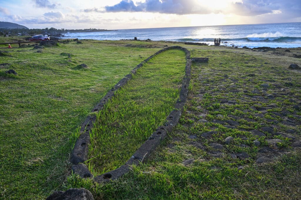 A grass field reaches towards the ocean at the top of the frame. The sun is setting at the top of the frame - sun rays and clouds above. On the grass is an elliptical shape set out in stones, its length reaching towards the ocean
