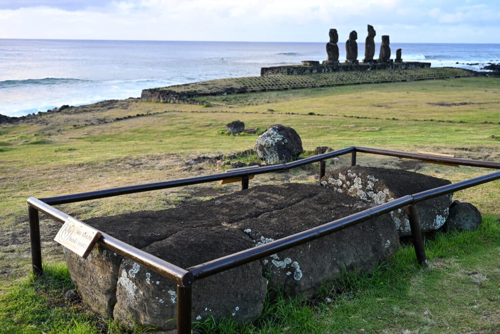 Looking down a grassy hill to the ocean beyond. In the foreground is a large carved stone statue lying on the ground with railings around it. In the distance is a wide stone platform with 5 stone statues upright