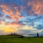 bottom third is green field. Top two thirds is a multi coloured sunset sky with clouds illuminated yellow, pink and orange light against a blue sky. A row of 5 stone statues are silhouetted to against the sky and a single statue to the right.