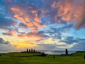 bottom third is green field. Top two thirds is a multi coloured sunset sky with clouds illuminated yellow, pink and orange light against a blue sky. A row of 5 stone statues are silhouetted to against the sky and a single statue to the right.