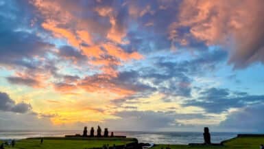 bottom third is green field. Top two thirds is a multi coloured sunset sky with clouds illuminated yellow, pink and orange light against a blue sky. A row of 5 stone statues are silhouetted to against the sky and a single statue to the right.