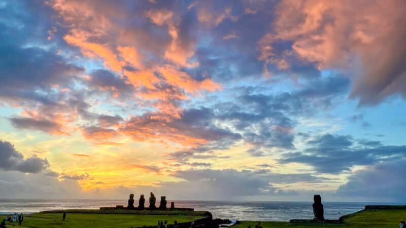 bottom third is green field. Top two thirds is a multi coloured sunset sky with clouds illuminated yellow, pink and orange light against a blue sky. A row of 5 stone statues are silhouetted to against the sky and a single statue to the right.