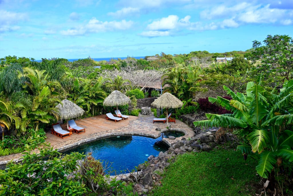 View looking down on a tropical garden with small swimming pool in the foreground surrounded by a row of small wooden huts and green foliage in background. Blue sky with small white clouds above