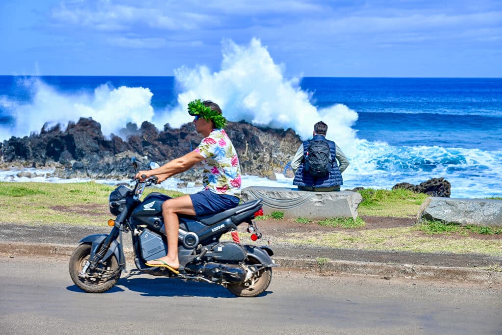 A man in flip flops, navy shorts, flowery Hawaii shirt and wreath of green leaves is driving on a motorbike from right to left of the frame. A man with backpack sits on coast behind watching waves crash over rocks