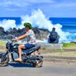 A man in flip flops, navy shorts, flowery Hawaii shirt and wreath of green leaves is driving on a motorbike from right to left of the frame. A man with backpack sits on coast behind watching waves crash over rocks