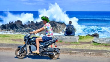 A man in flip flops, navy shorts, flowery Hawaii shirt and wreath of green leaves is driving on a motorbike from right to left of the frame. A man with backpack sits on coast behind watching waves crash over rocks