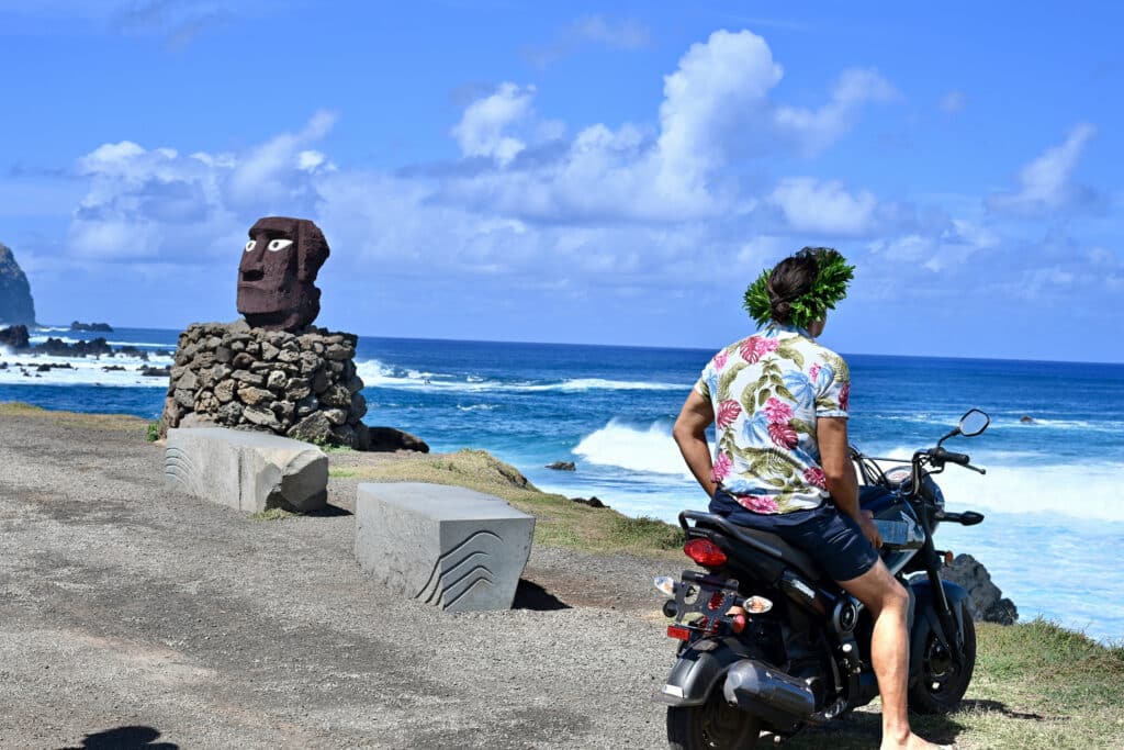 A man wearing a Hawaiian shirt sits on a moped looking out to the ocean. He has a wreath of green leaves on his head. To the left is a statue of a head with white eyes in dark stone blue sky above the ocean