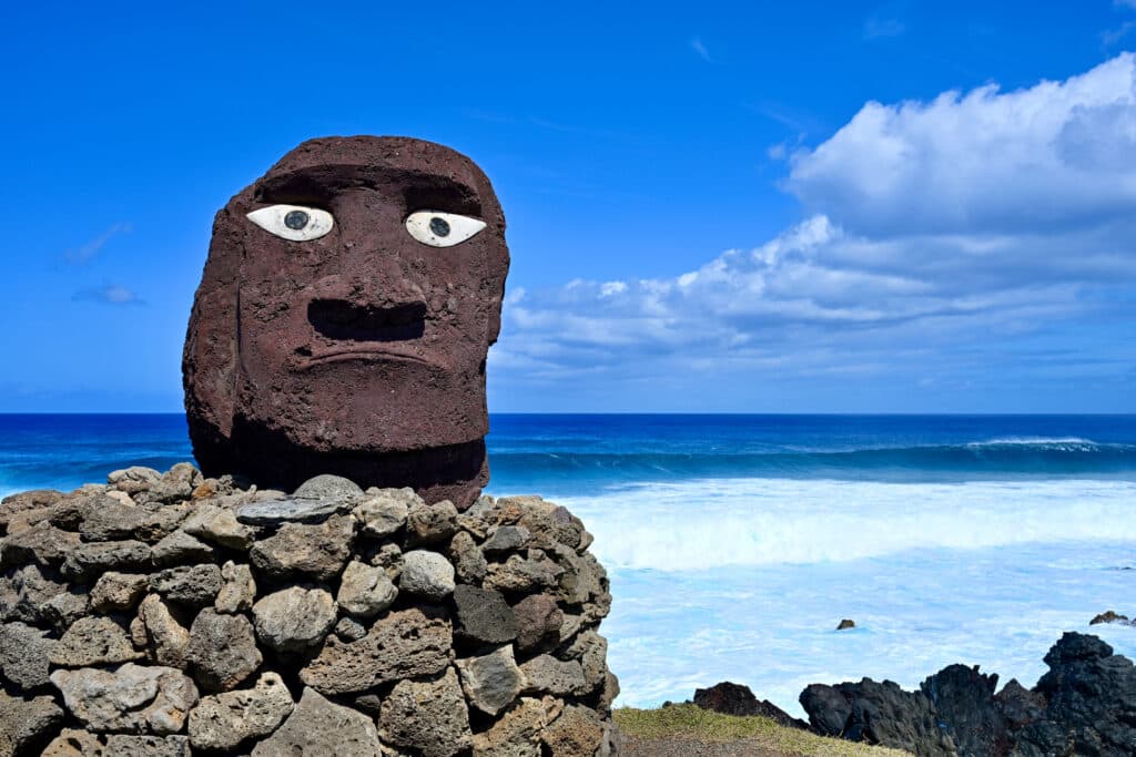 A large stone head in dark rock sits on a pile of stones to the left of the frame. White eyes. Turquoise ocean to the right hand side. Blue sky above.