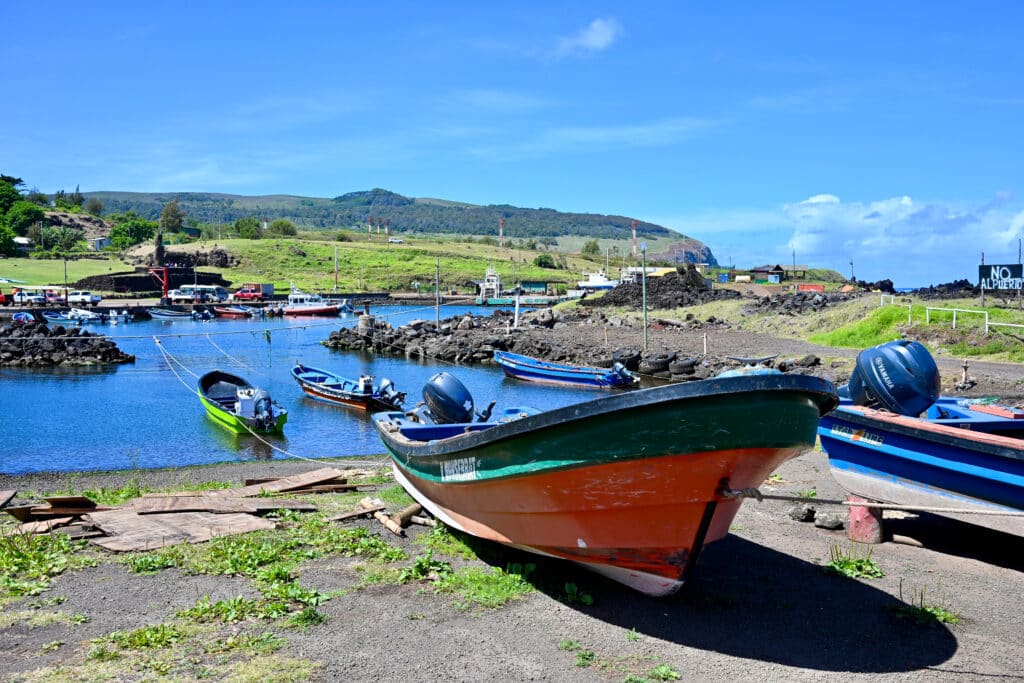 View of a small red and black fishing boat moored on the right hand side with a small harbour behind it and hills on the horizon. Blue sky