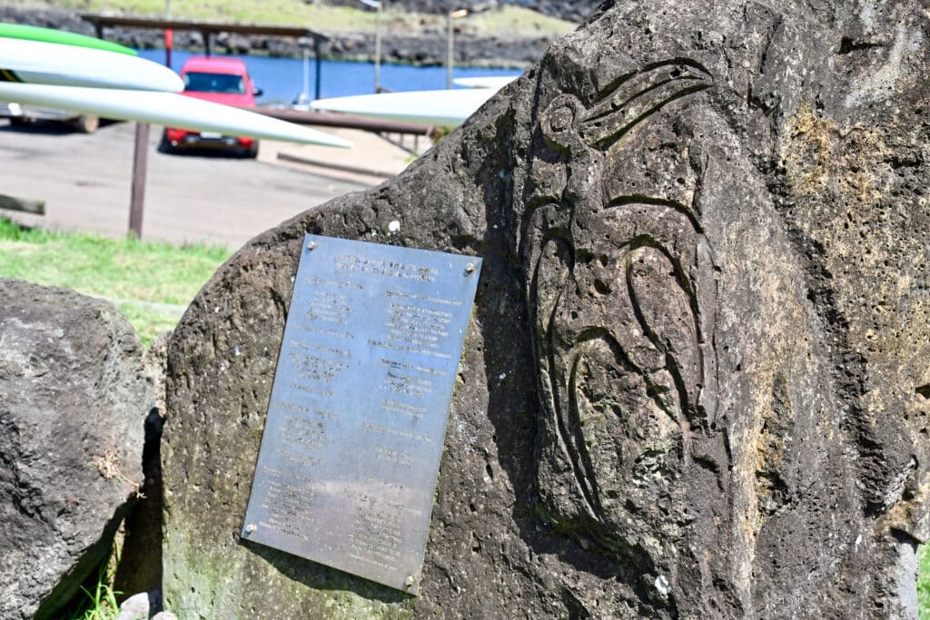 A dark stone rock featuring the carving of an upright bird figure on the right hand side. Harbour behind to the top left