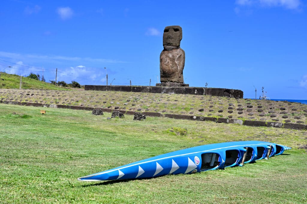 A long thin blue canoe lies on its side to the right on a patch of pale green grass. a wide stone platform runs across the horizon with a single stone statue with large head and torso. Blue sky behind