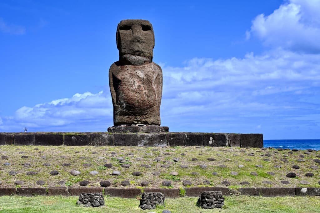 View of a large human form statue with large head and torso standing on a stone platform on grassy hill scattered with stones. Blue sky behind