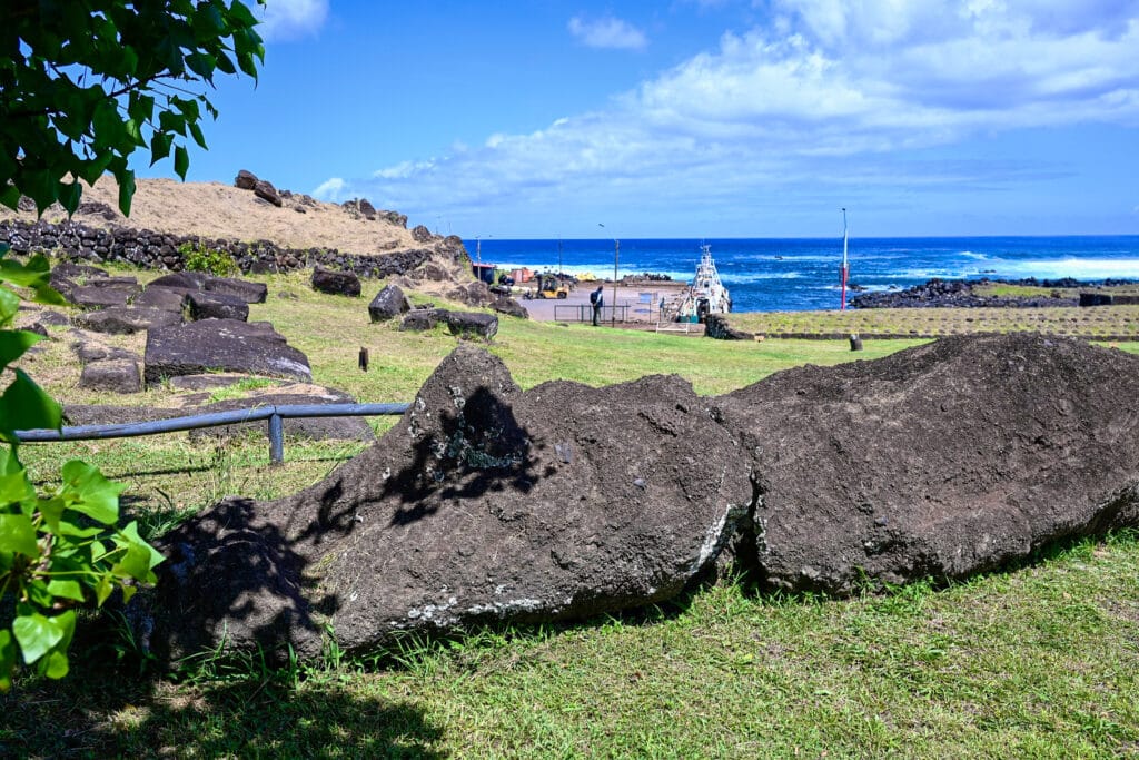 A human form statue is lying face up on a grassy hillside with ocean view in the background