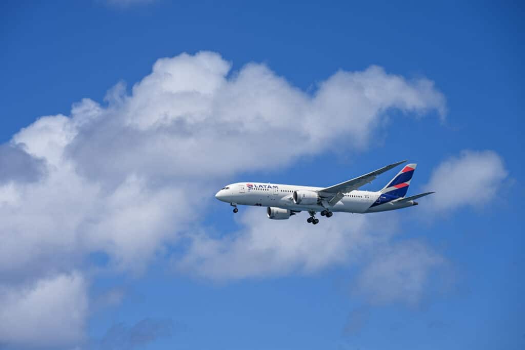 Blue sky with big white fluffy clouds across the middle. A white airplane is flying into the frame from the right hand side. It has LATAM log on it