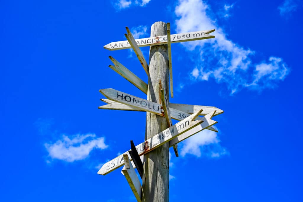 Blue sky with a wooden post in the front supporting pale wooden arrows pointing to various locations such as San Francisco, Honolulu, with the distances marked on