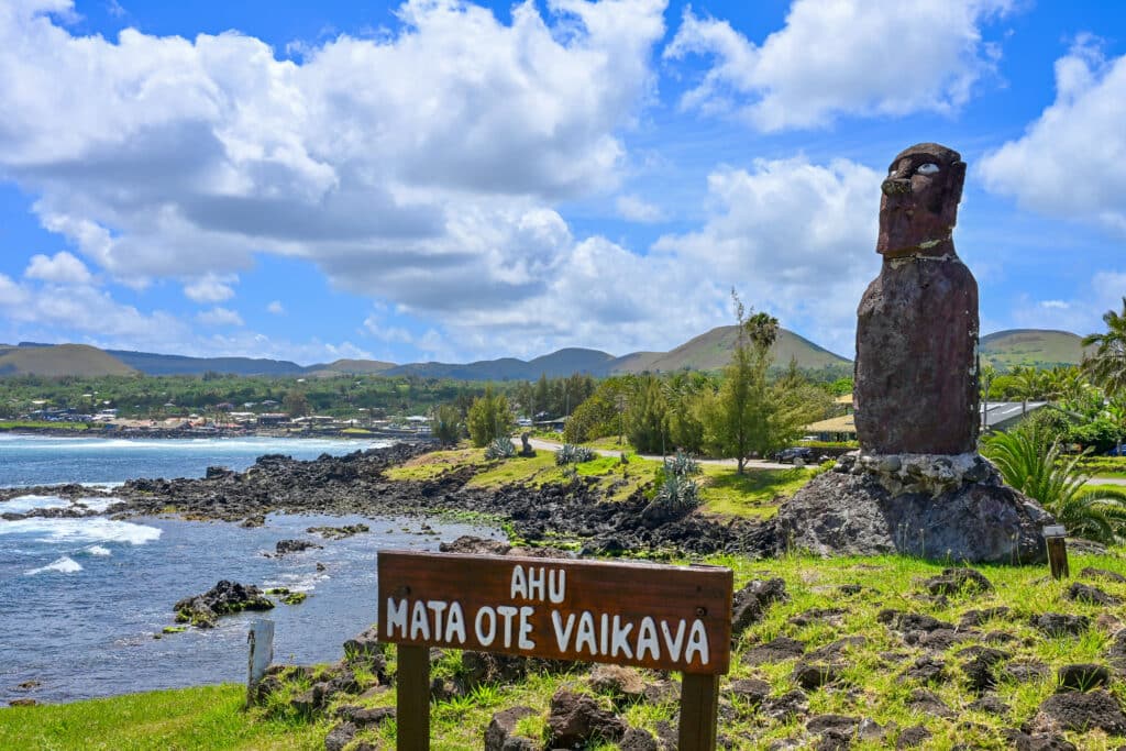 A large stone statue (moai) standing on a small rocky platform stands to the right hand side of the frame on a rugged coast. Ocean to the left. Hills in the background. Cloudy blue sky above. Wooden sign saying 'Ahu Mata One Vaikava' at the bottom of frame