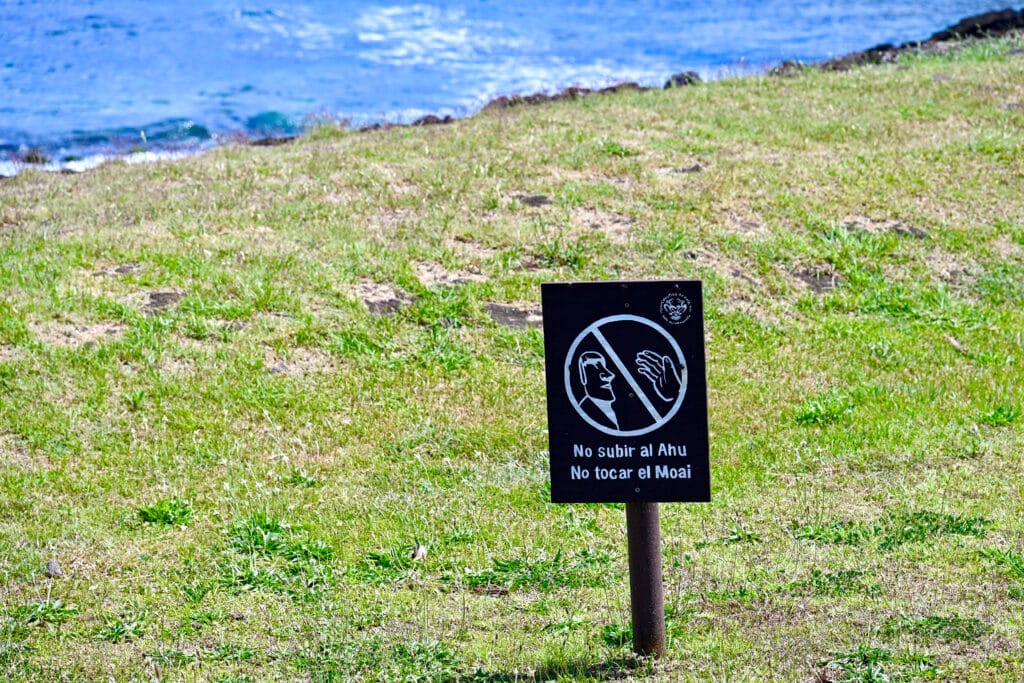 A grass bank in the foreground with ocean visible in top left corner. A rectangular wooden sign sits on the grass and features a white design to tall you not to touch the statues