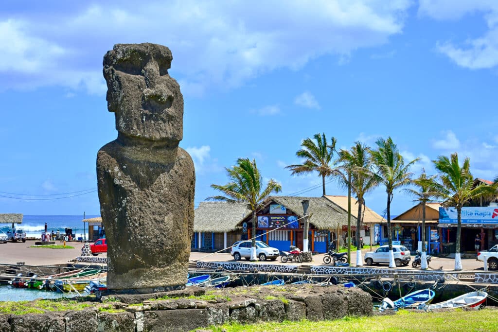 A large stone statue in human form stands to the left hand side. Palm trees and low rise building to the right. Pale blue sky above