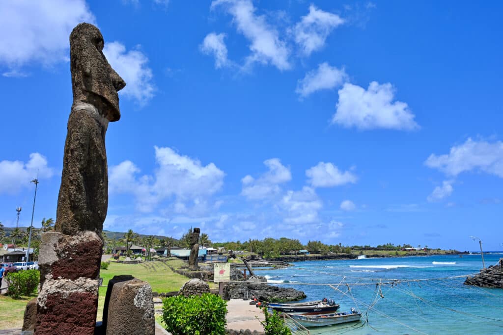 A single moai stands to the left hand side overlooking the ocean with a grassy coastline in the background and blue sky above