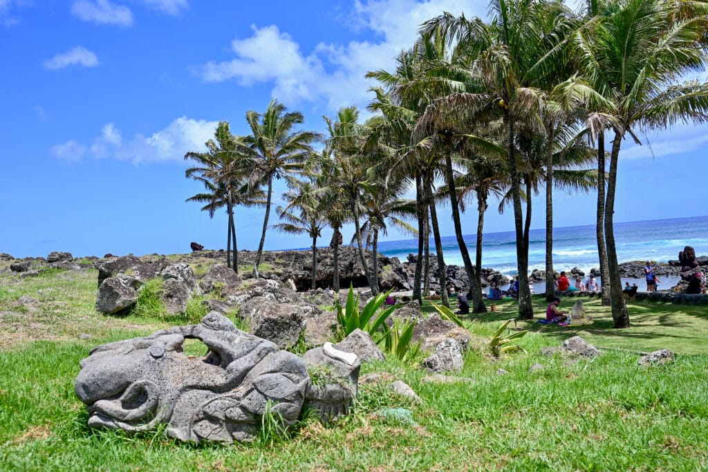 Carved stone sits to the bottom left of the frame on grass with row of palm trees to the right hand side. Ocean behind and blue sky above