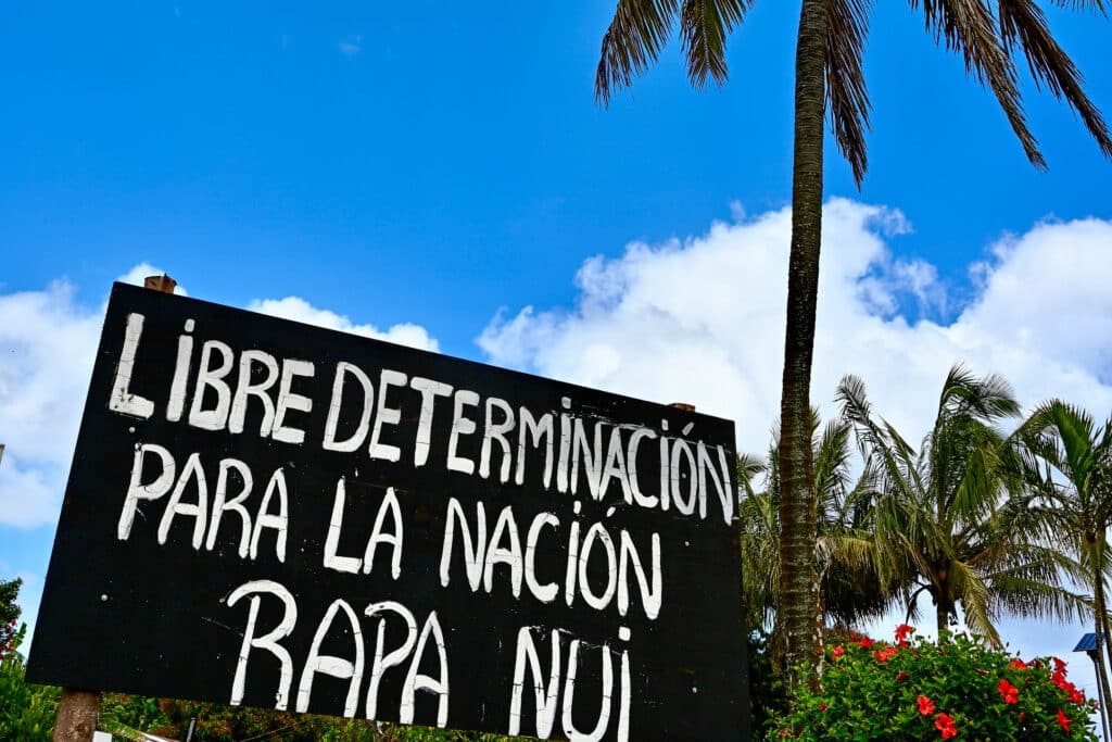Looking up at a black sign in the bottom left hand side of the frame. It has white letters in Spanish - a slogan about liberation for Rapa Nui. Blue sky behind. Palm tree and tropical plants to the right hand side