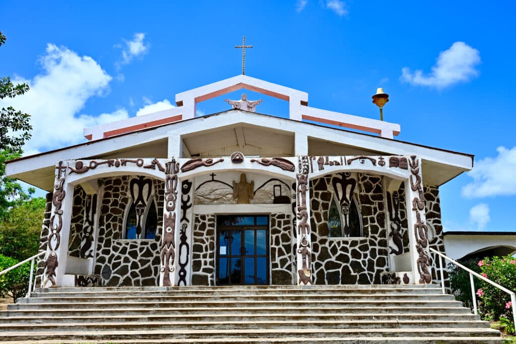 Stunning outside of a white church building. Photo looking up stone steps to the front of the church. Exterior of church is covered with brown Polynesian motifs and designed. A Christian Jesus figure on the roof and cross on top. Tropical flowers and greenery to the sides