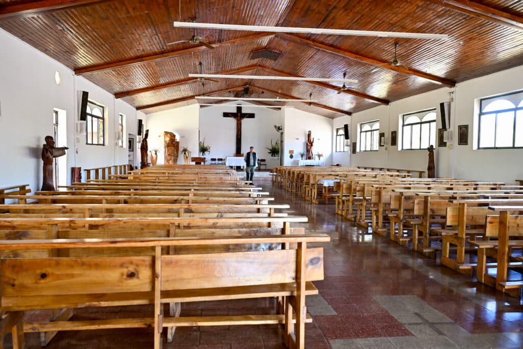 Inside of a simple church with white walls but wooden ceiling and wooden pews throughout. Wooden statues and carvings around the room. A man is standing at the far end of the church. red and grey tiled floor