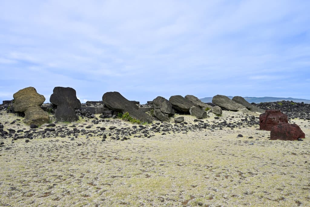 Pale green grassy field with row of large stones (could be statues) lying across the middle. Pale blue cloudy sky above
