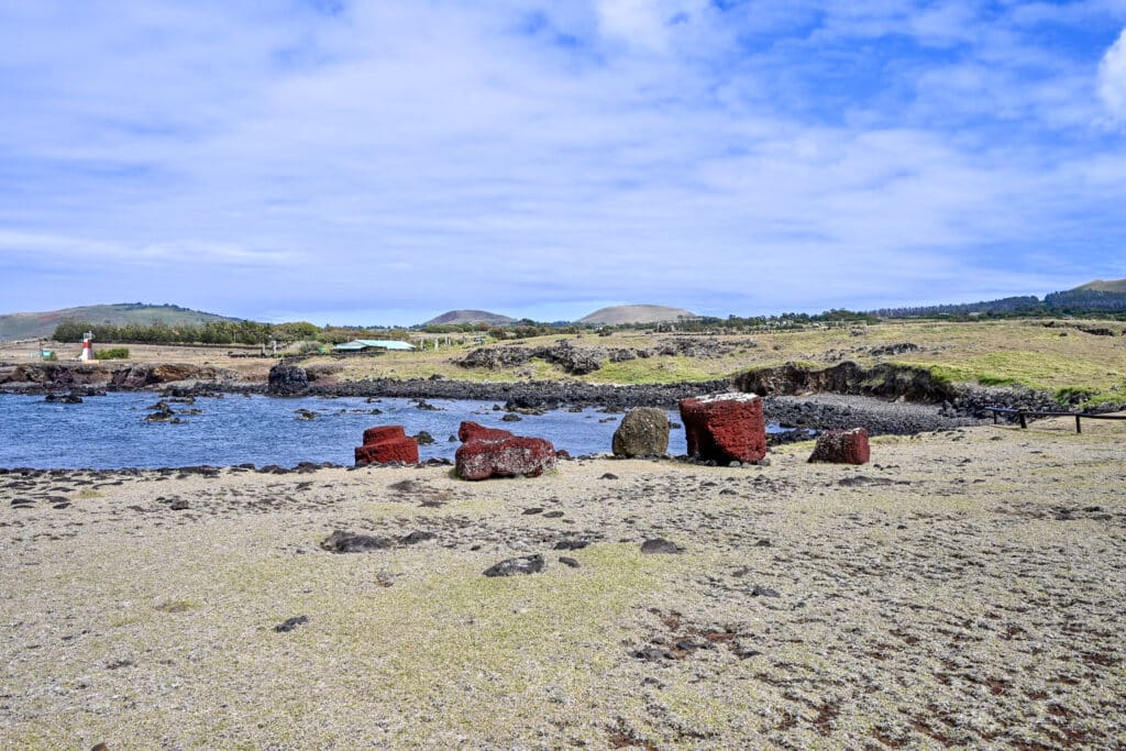 Pale yellowy green grass in foreground with water to the left with blue sky above. A number of red circular stones lie on the edge of the water
