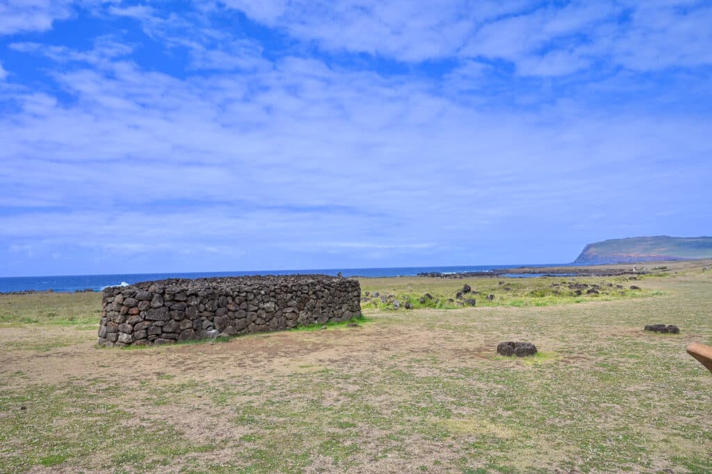 A field of patchy green grass leads to the ocean with cloudy blue sky above. In the middle is a long and low structure made of dry stone blocks