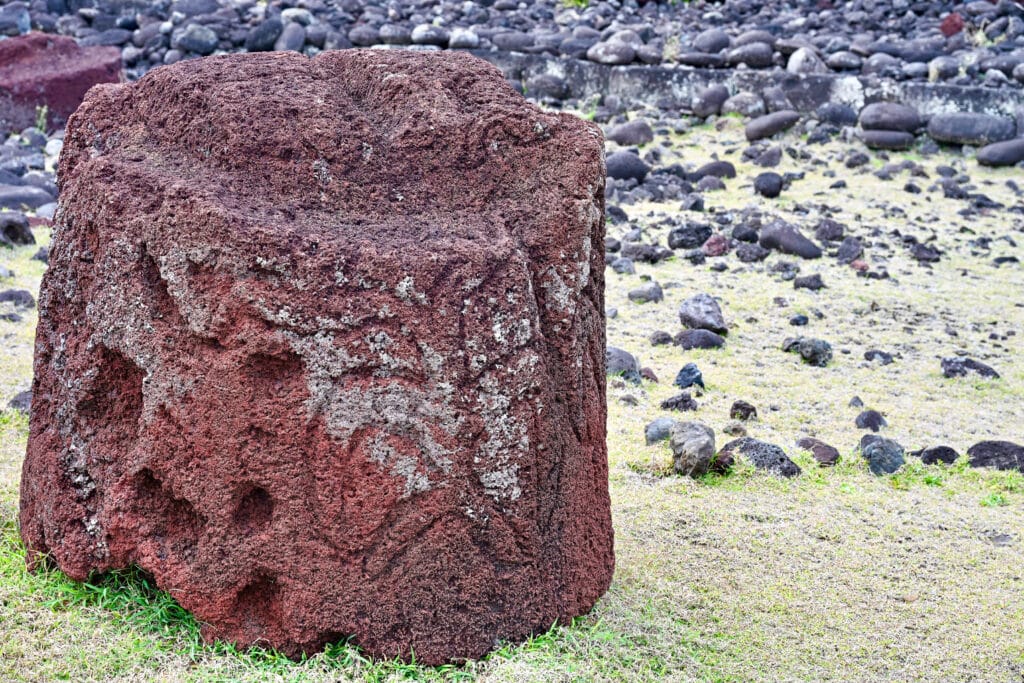 A circular red stone sits on pale yellow grass to the left. It has carvings on the stone. Black stones in the background